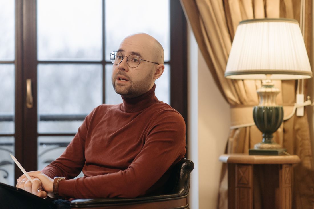 A man in a rust-colored sweater sits mid-conversation in a leather armchair, lit by soft daylight from a window and warm lamplight in a plain room.
