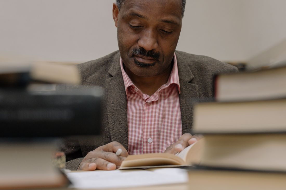 Man in a blazer reading among stacks of books, representing the scholarly setting behind a faculty portrait