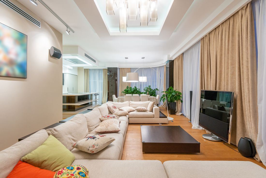 Spacious modern living room with beige sectional sofa, wall-mounted television, and floor-to-ceiling curtains