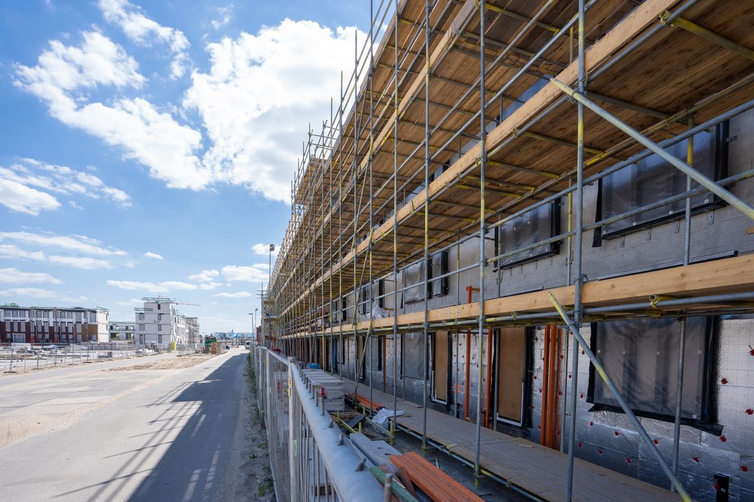 A multi-story building under construction, its concrete facade wrapped in steel scaffolding and plywood decking, seen from a street running alongside the site under a partly cloudy blue sky.