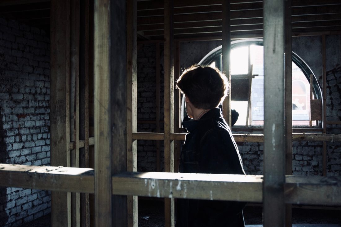 A person walks through the exposed wooden framing of a building's interior, backlit by light from an arched window opening, with brick walls visible on either side.