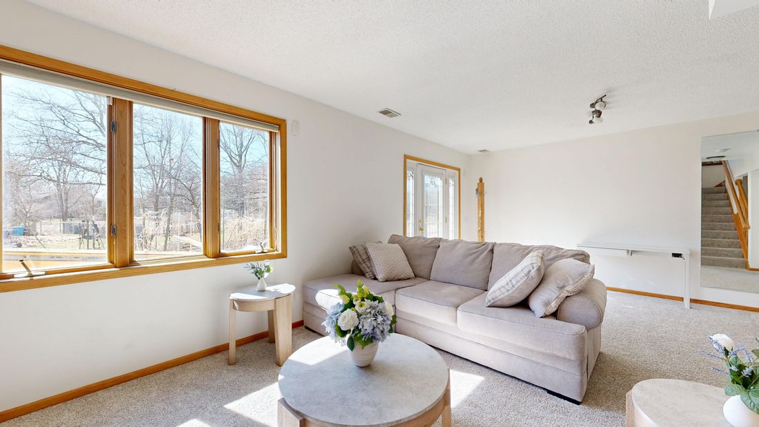 Empty, staged living room with a beige sectional sofa, fresh flowers on a side table, and large windows letting in bright natural light.
