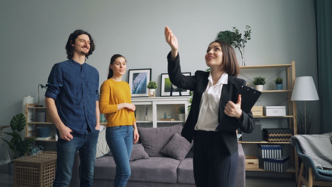 A small group tours a bright, furnished apartment living room together, taken from a distance with the couch, shelving, and plants visible.