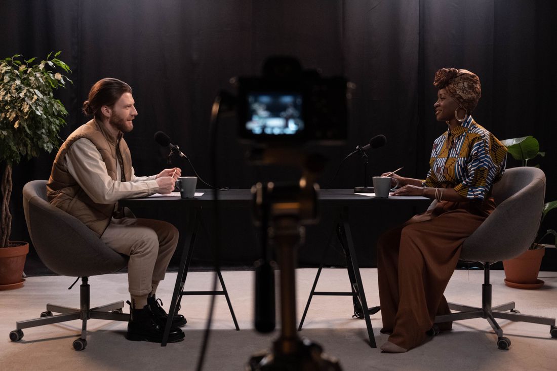 A video interview being filmed in a studio, two people talking across a desk with a camera and microphones set up between them.