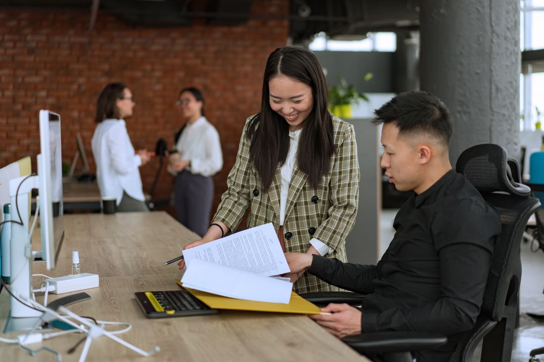 Two coworkers reviewing documents together at a desk in an open office, with colleagues talking in the background.