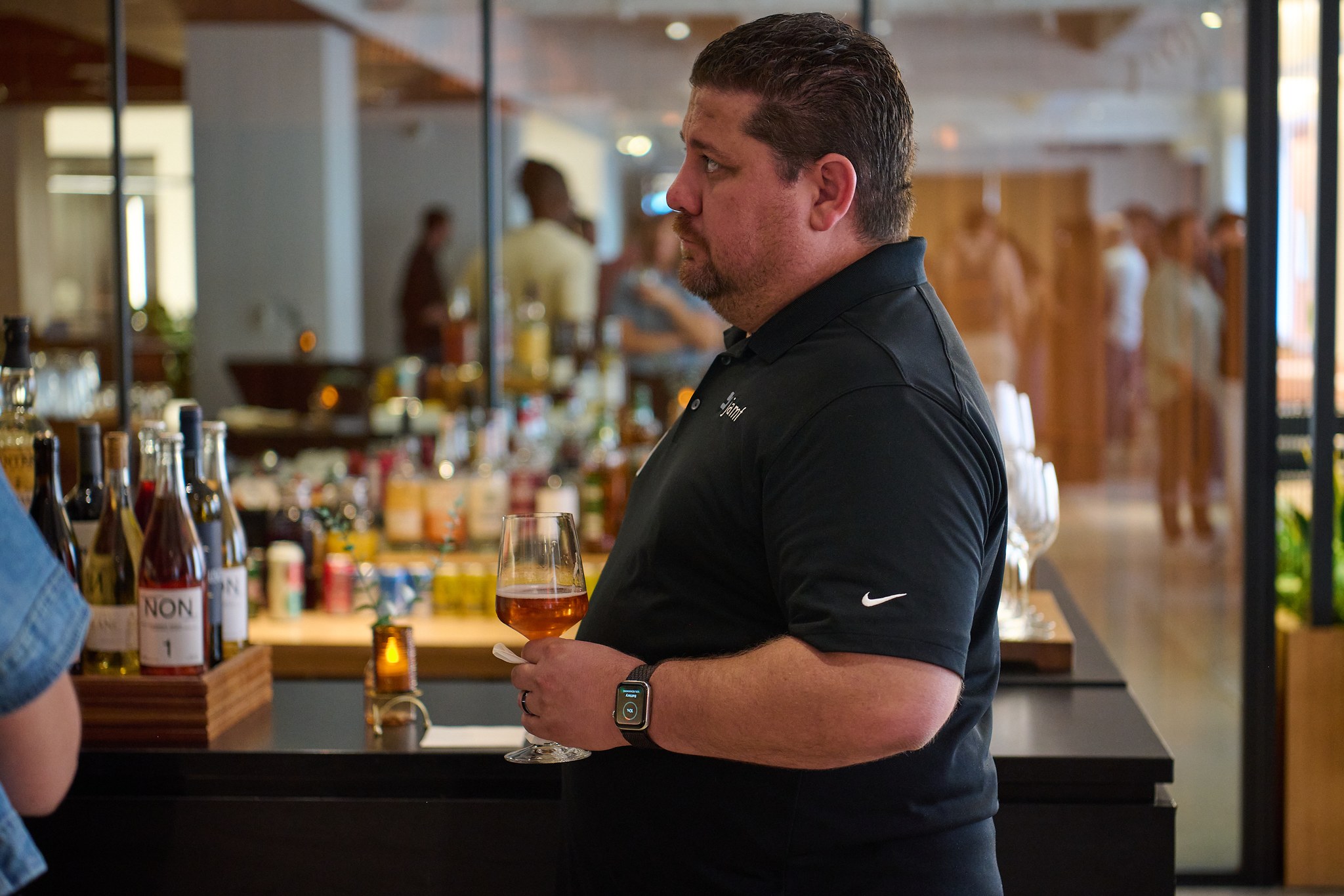 Man in polo shirt at event bar holding wine glass with people in background.