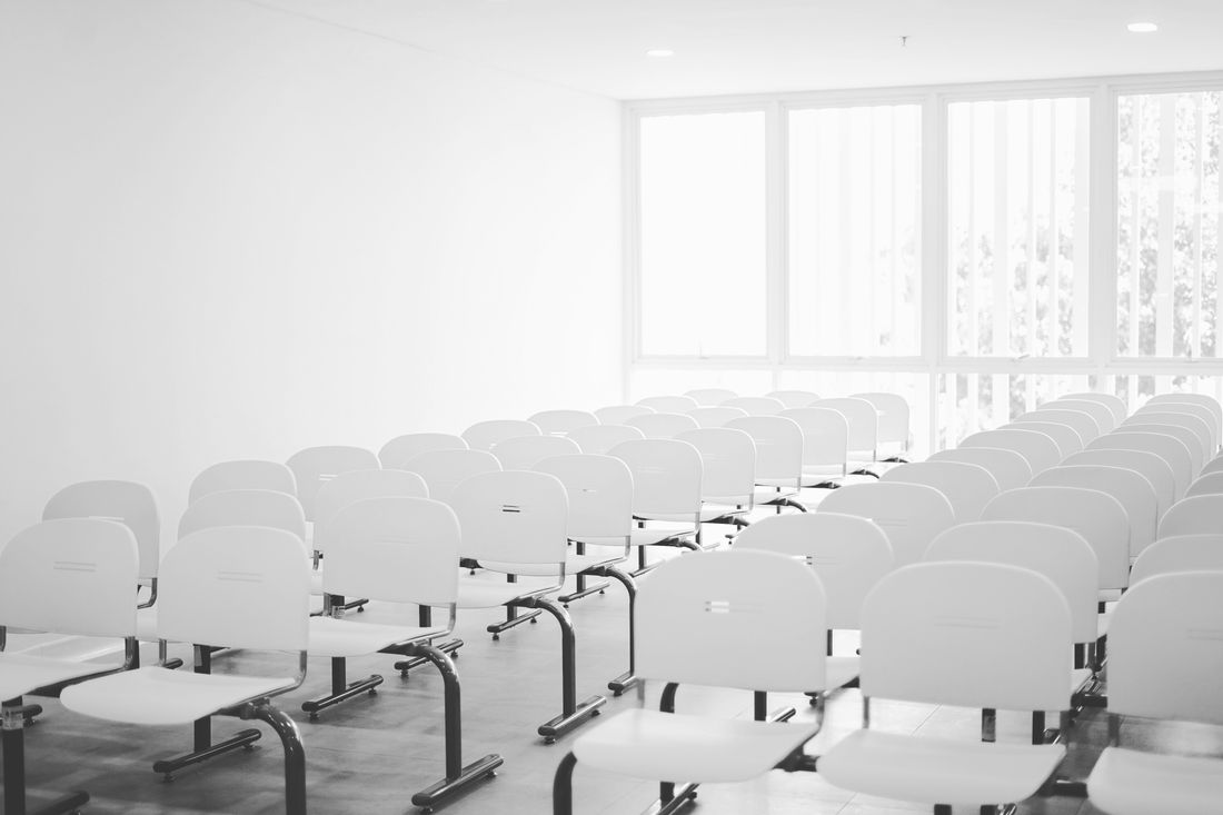 Rows of empty white chairs face a bright bank of windows in a plain, unbranded meeting room before an event begins.