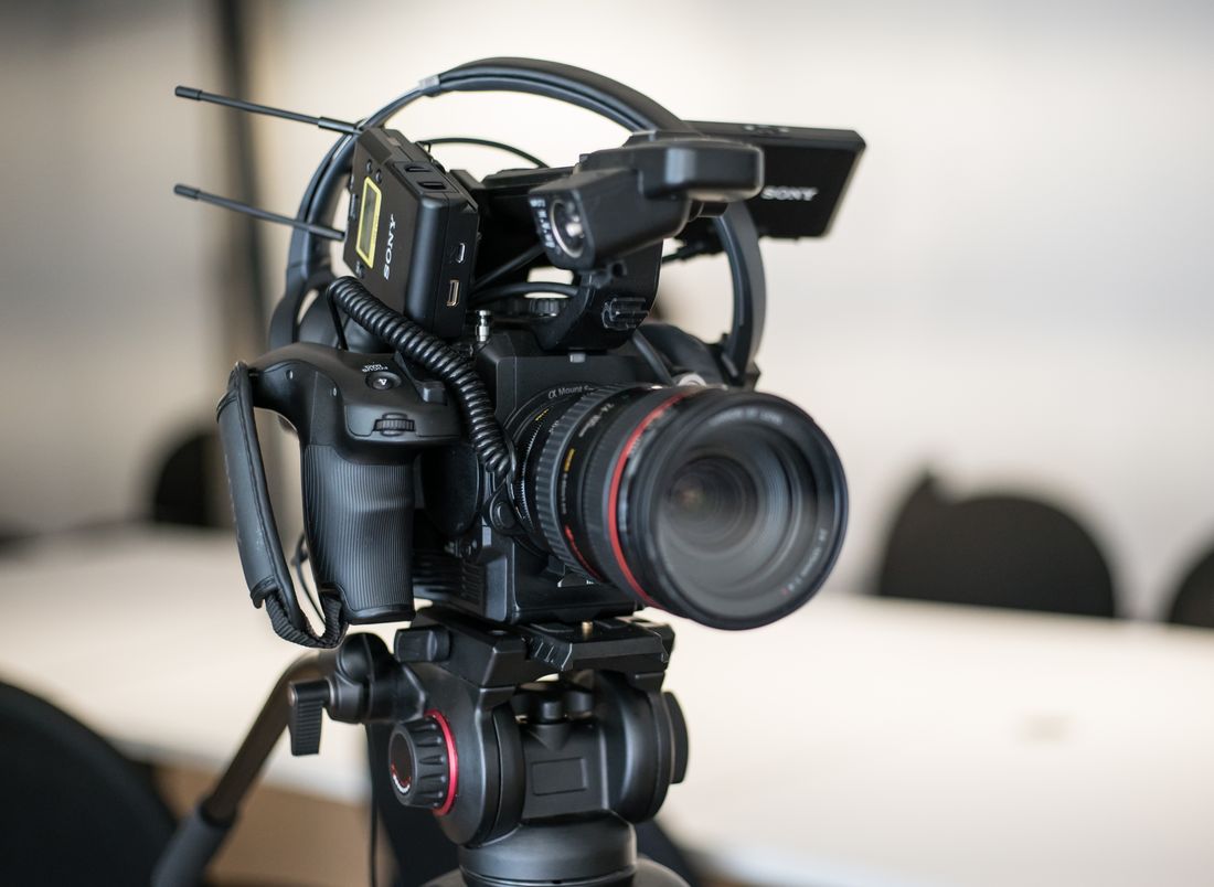 A professional video camera on a tripod stands in sharp focus in front of blurred tables and chairs in a plain meeting room.