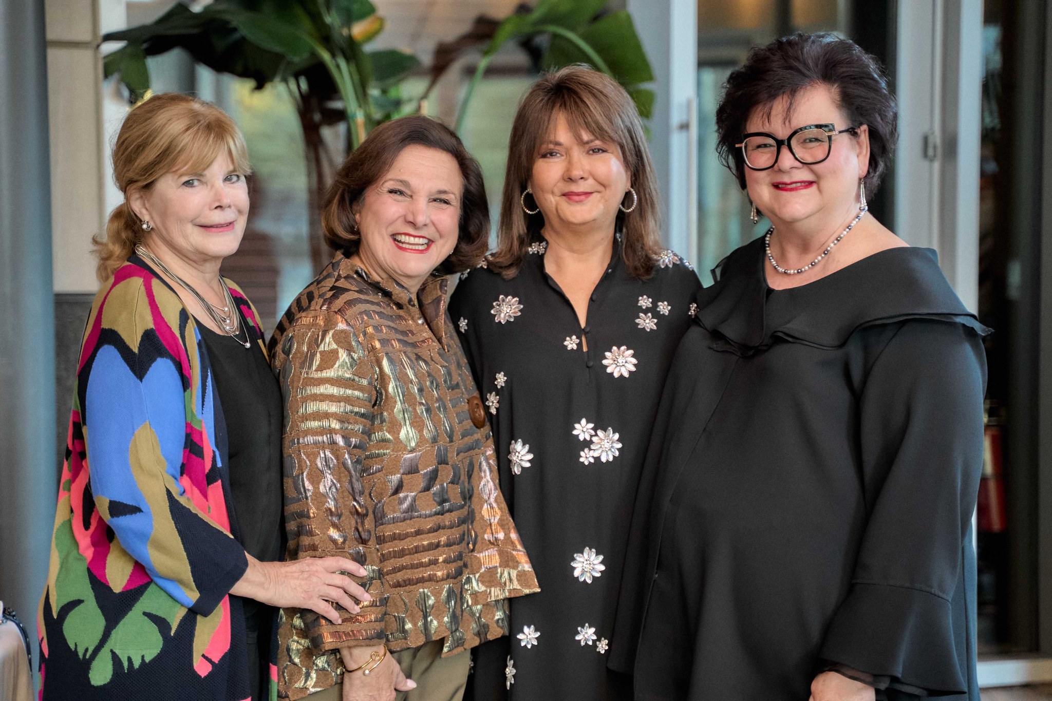 Four women in dressy outfits posed together smiling indoors, celebration.