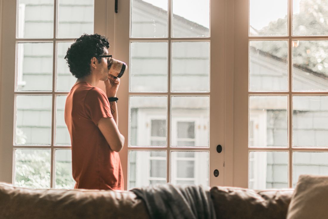 A person in a plain orange t-shirt stands in profile near a bright apartment window, holding a mug, with a couch in the foreground.