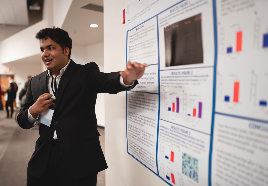 A presenter gestures toward data on a printed research poster while explaining results in a conference hallway.