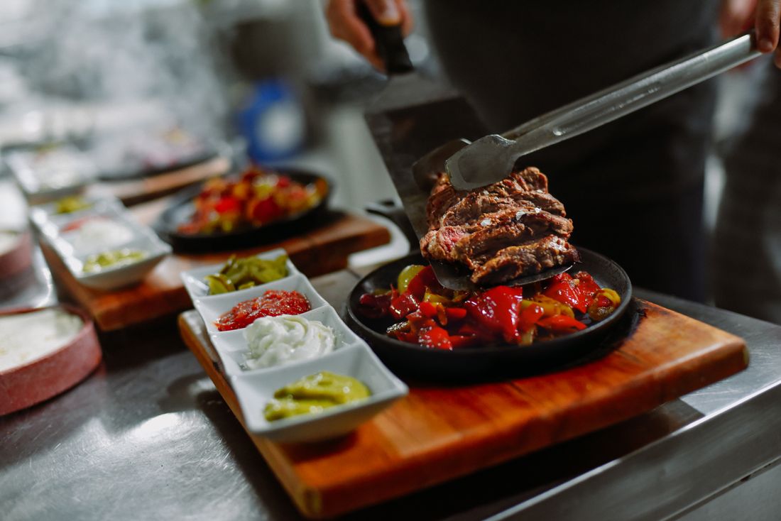 A sizzling skirt steak fajita plated with tongs onto a hot cast-iron dish alongside small bowls of salsa, sour cream, and guacamole, a kitchen counter blurred behind it.