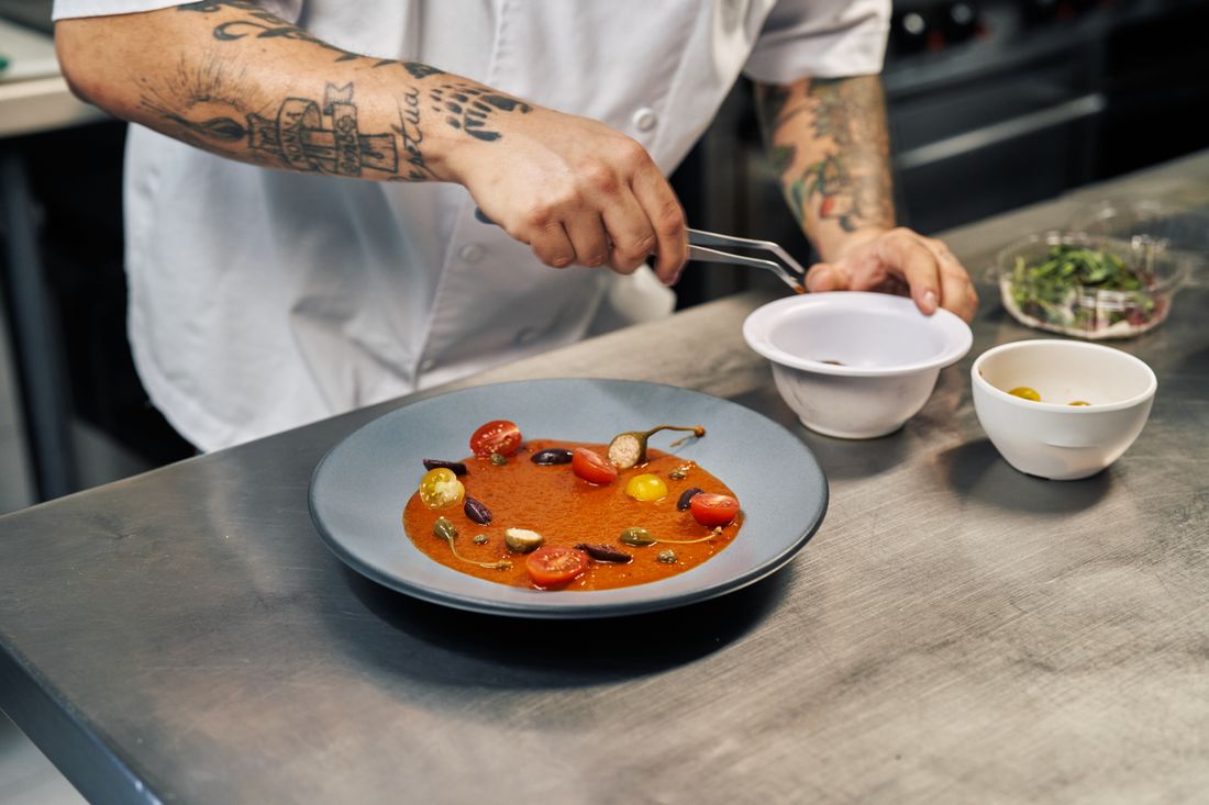 A cook's tattooed hands use tweezers to place a cherry tomato garnish on a plated tomato dish at a stainless steel kitchen counter, small prep bowls nearby.