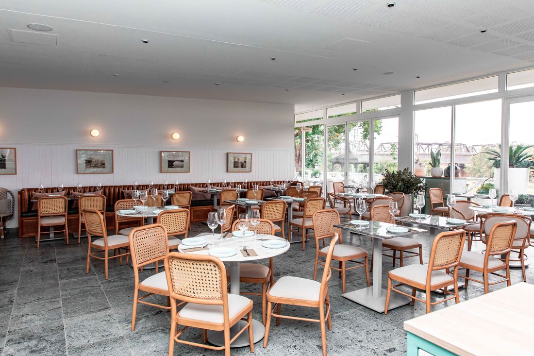 An empty, sunlit restaurant dining room with cane chairs and set tables, natural light streaming through floor-to-ceiling windows before service begins.