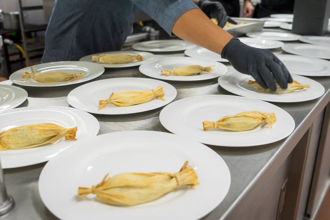 A gloved kitchen hand places a plated tamale onto one of many white plates lined up along a stainless steel counter during service prep.