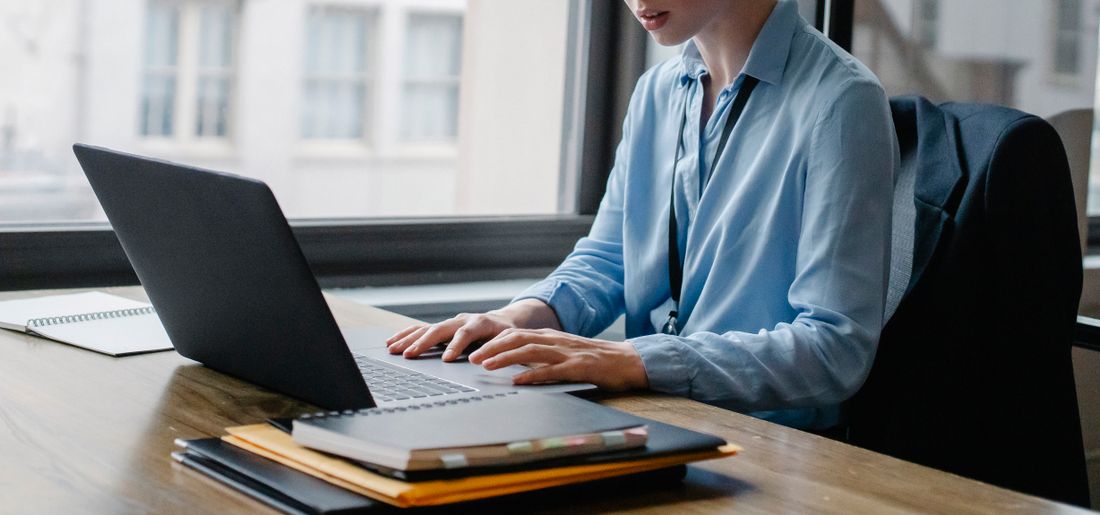A professional at a wooden desk works on a laptop near a window, with a notebook and folders stacked nearby and a blazer on the chair behind her.