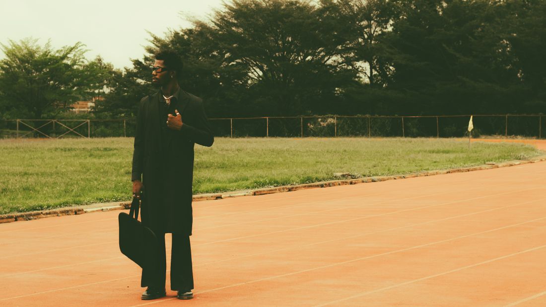 A man in a long dark coat holds a briefcase while standing on an outdoor running track beside an open field.