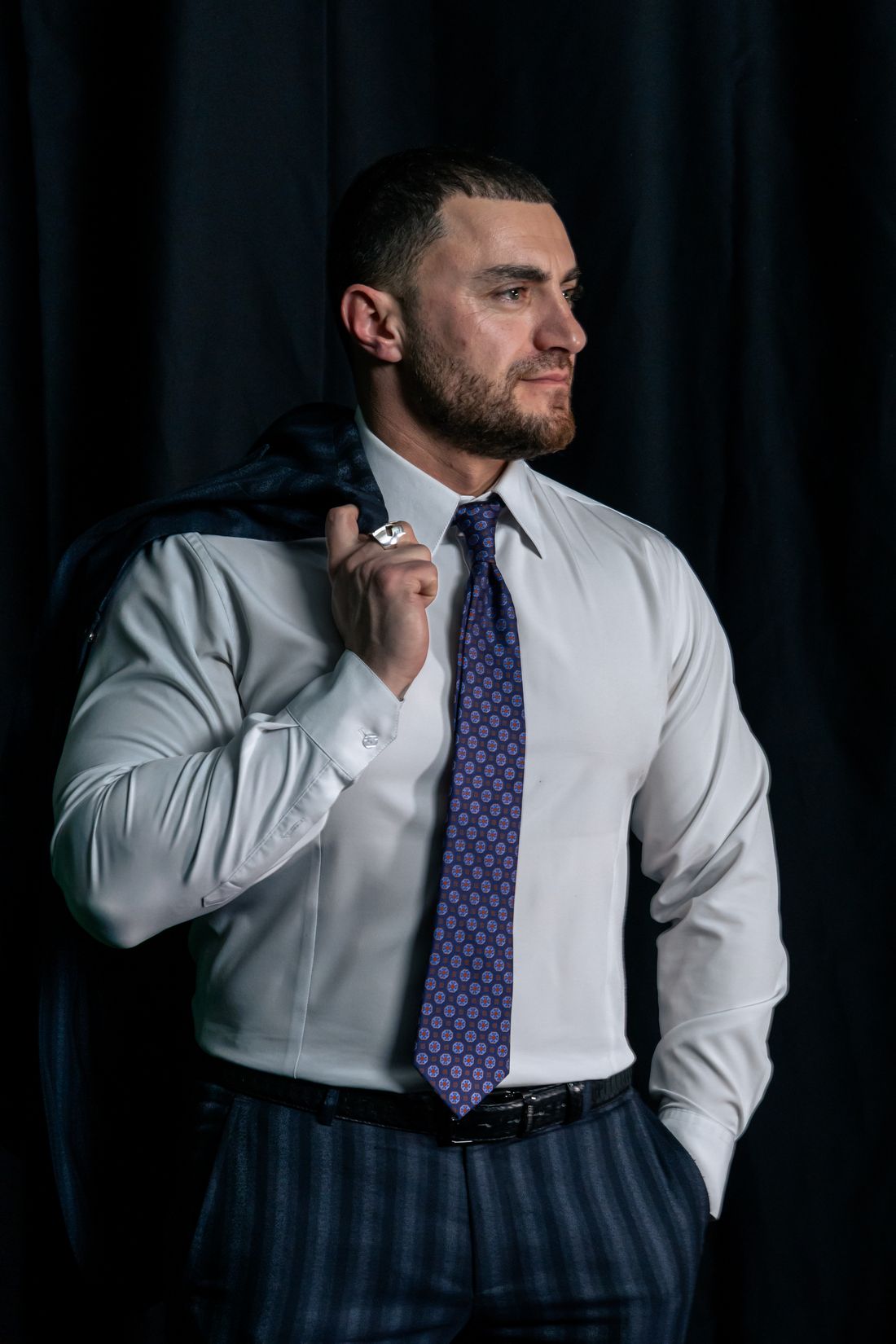 A muscular, athletic-built man in a white dress shirt and patterned tie holds his suit jacket over one shoulder, posing against a plain dark background.