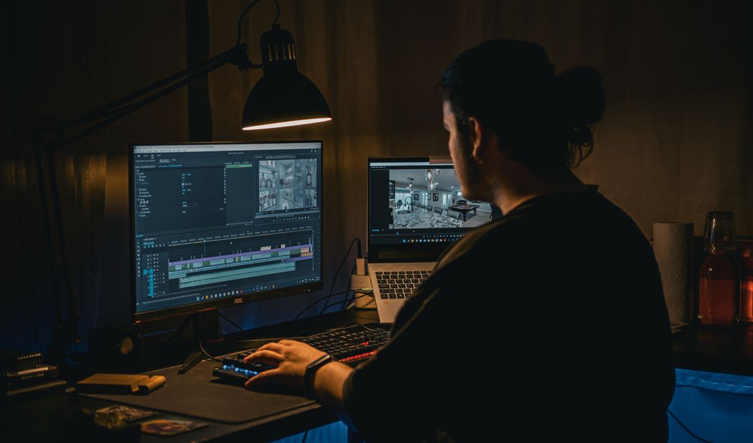 A video editor working late at a dual-monitor setup in a dimly lit room, desk lamp lighting the timeline on screen