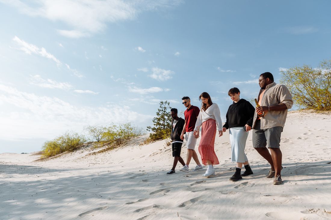 A small group of five colleagues in casual clothes walk together along a sunlit beach, mid-conversation, seen from a slight distance.