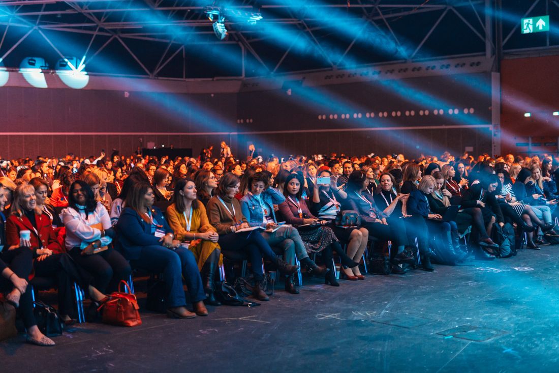 A large seated audience with conference lanyards watches a presentation under dramatic blue stage lighting.