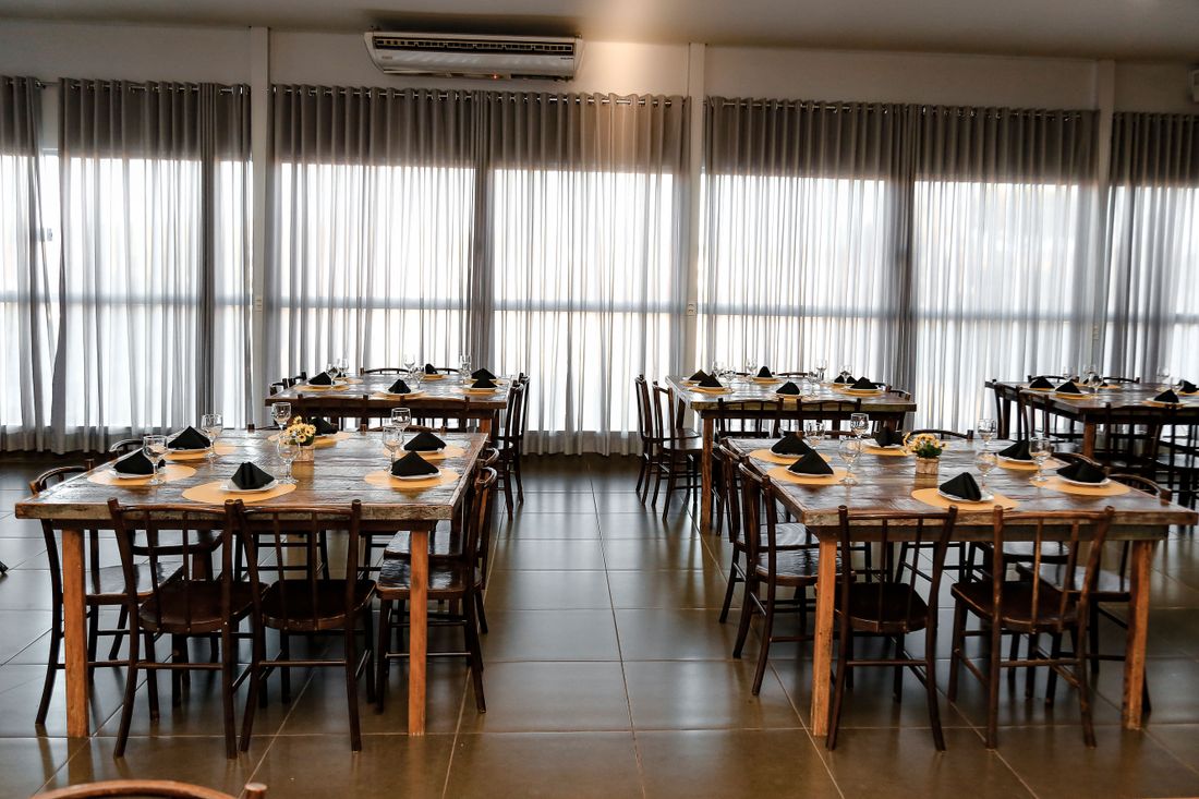 An empty event dining room with wooden tables set with black napkins and place settings, sheer curtains along the windows, no guests yet.