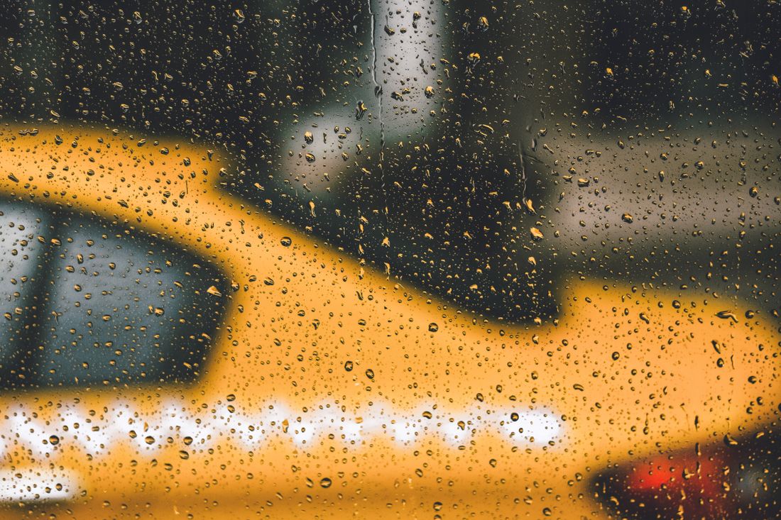 Midtown traffic and a yellow taxi seen through a rain-streaked car window, everything softened by water droplets and motion.