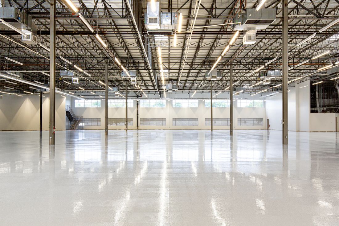 An empty industrial space with a polished concrete floor, exposed overhead beams, and no people, lit by daylight from a row of high windows.