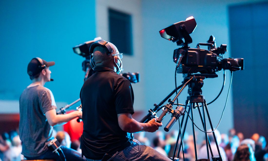 Two camera operators positioned side by side filming a stage at an indoor event, each on a separate camera