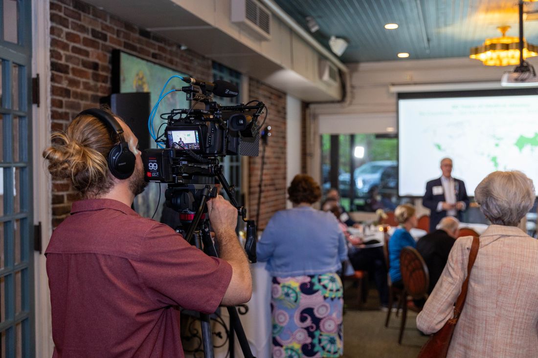 A videographer filming a corporate conference session with attendees seated and a presentation screen visible in the room