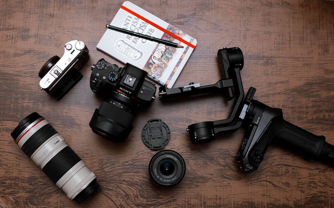 An overhead view of camera bodies, a telephoto lens, a gimbal stabilizer, and a notebook laid out on a wooden table.
