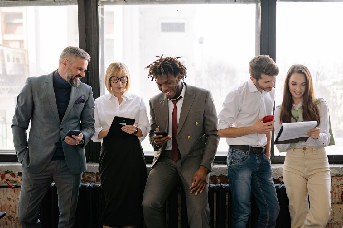 Five colleagues in business attire stand together by a bright office window, smiling and checking phones and papers.