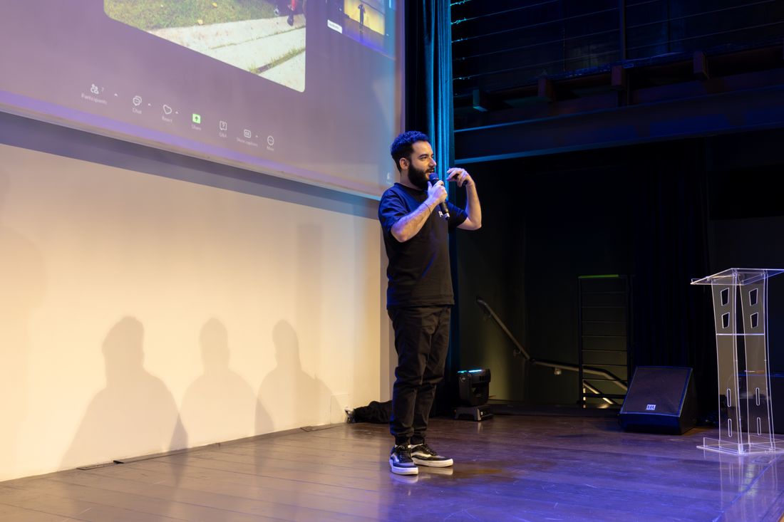 A speaker holding a microphone mid-talk at a corporate event, with presentation slides projected behind him.