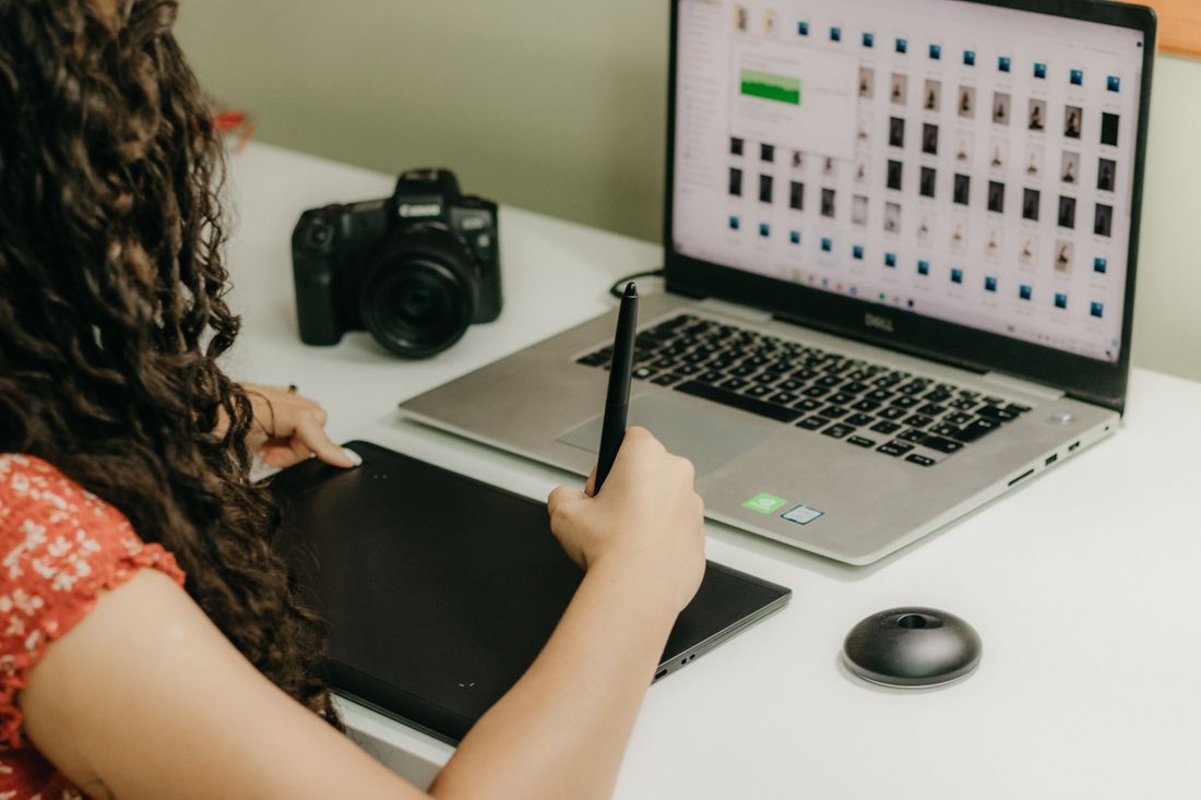 A person reviewing a grid of photo thumbnails on a laptop screen next to a camera and a graphics tablet.
