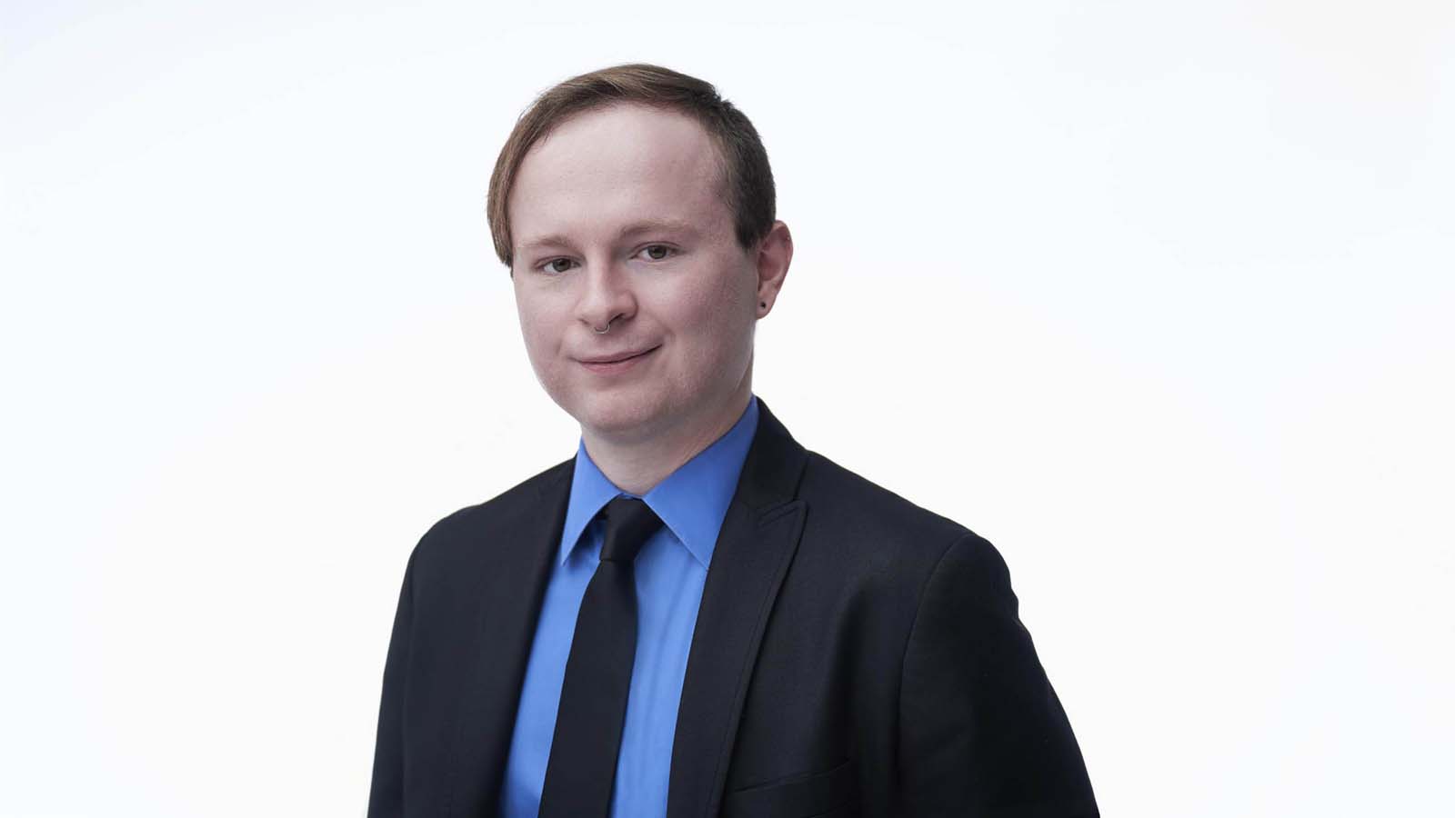 A young professional with a nose ring photographed at a slight three-quarter turn in a blue shirt and black suit against a plain white background