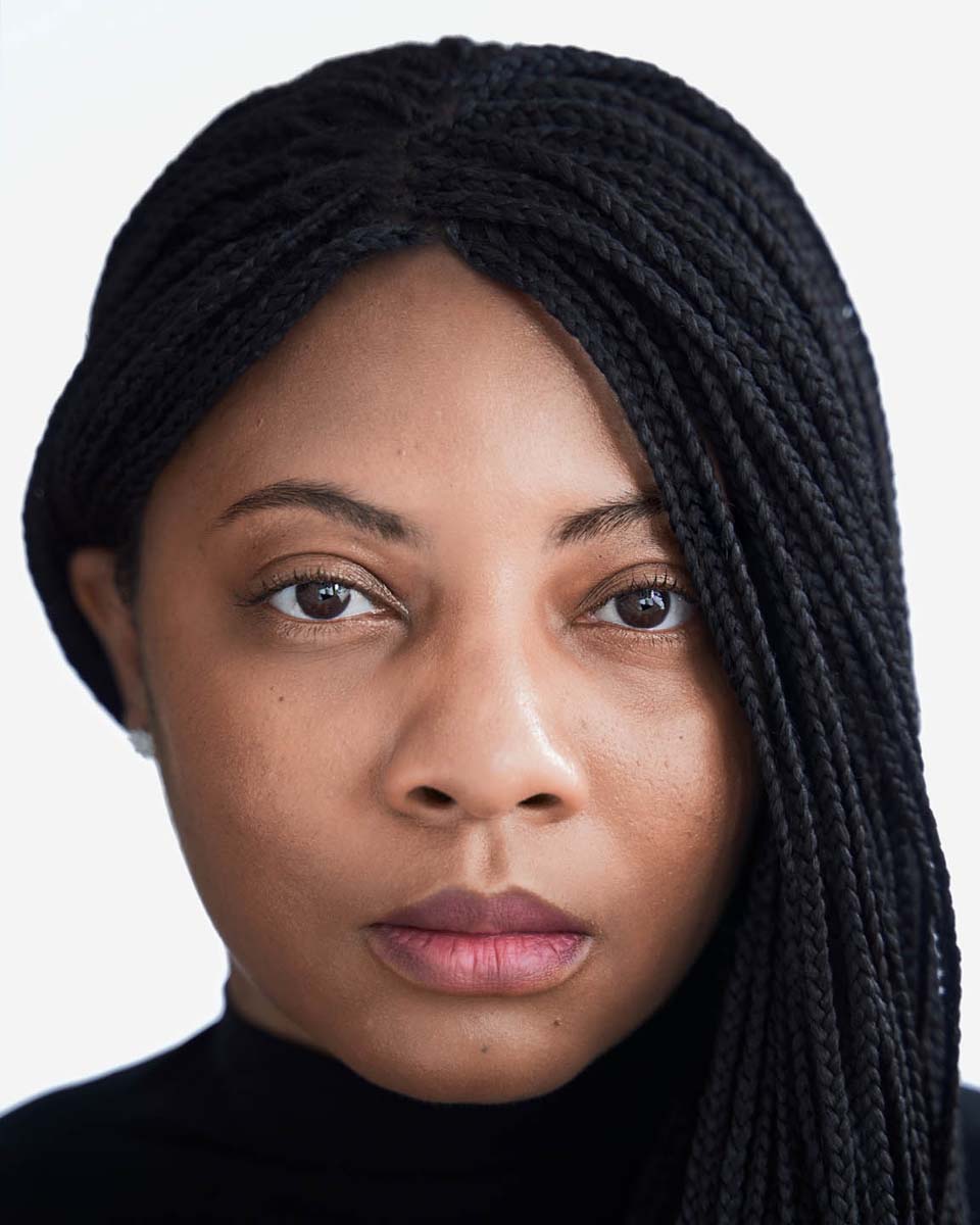 A close, tightly cropped portrait of a woman with braided hair in a black turtleneck against a plain light background
