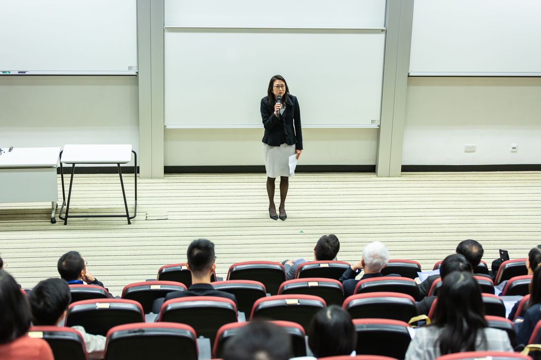A speaker standing at the front of a bright lecture room facing rows of seated attendees
