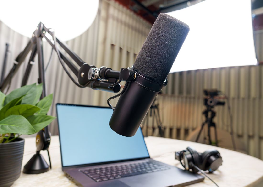 A podcast microphone on a boom arm beside an open laptop and headphones on a desk