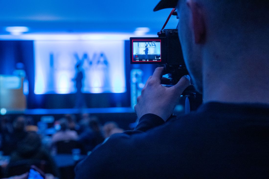 A camera operator films a speaker presenting on stage at a conference, framing the shot on the camera's flip screen.