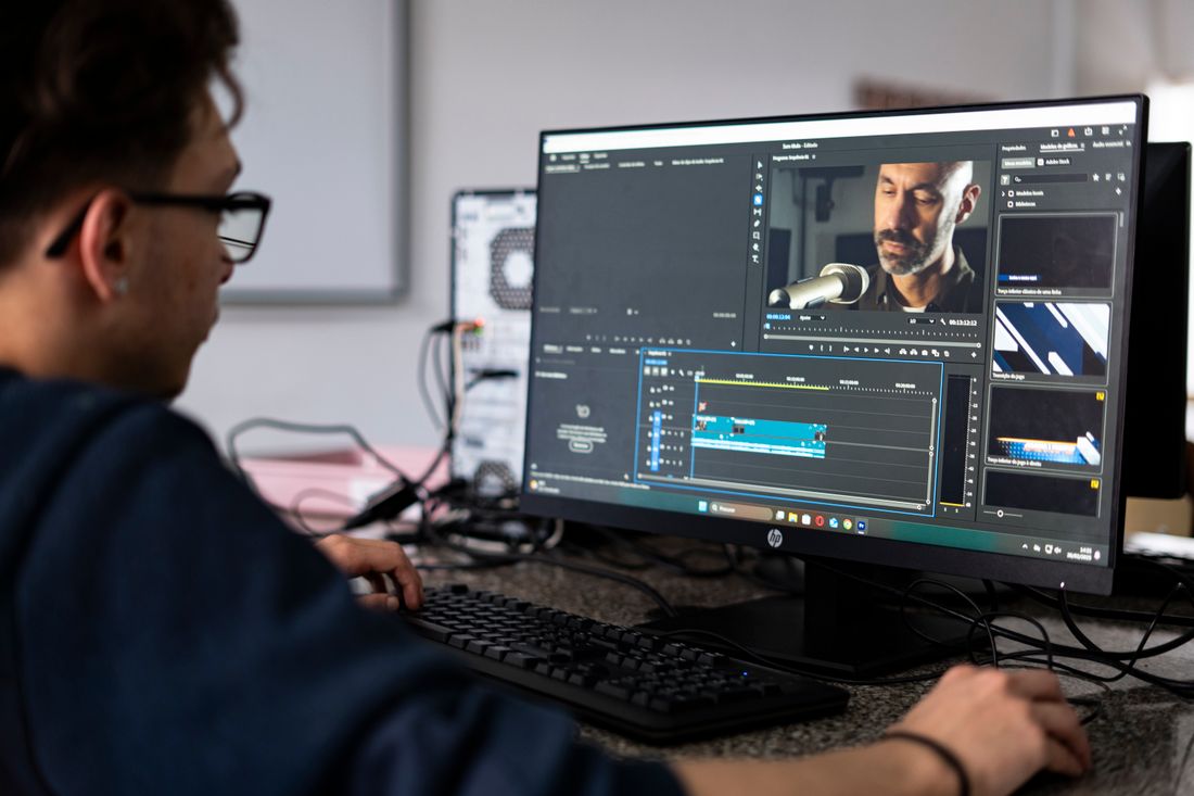 A video editor at a dual-monitor setup cuts together talking-head footage of a speaker on a timeline.