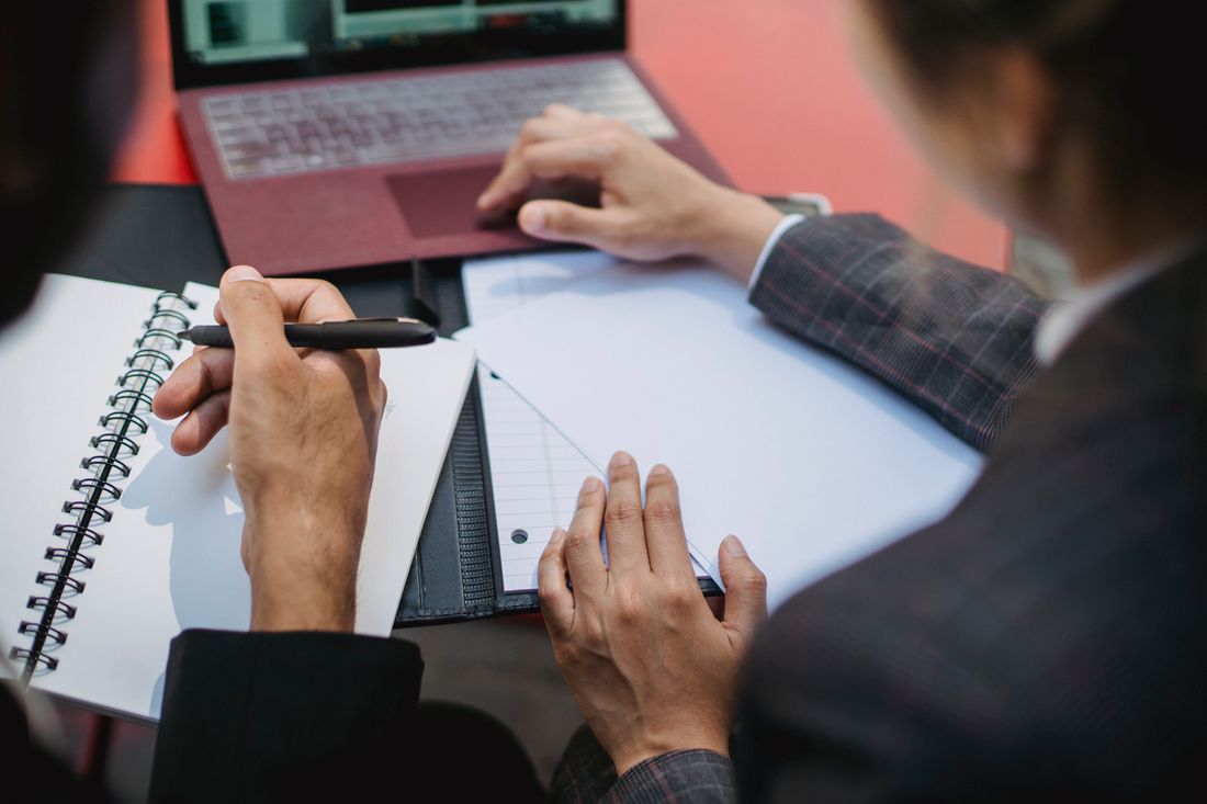 Close, over-the-shoulder view of two people's hands over a blank notebook and blank ruled paper on a desk, with an open laptop nearby.