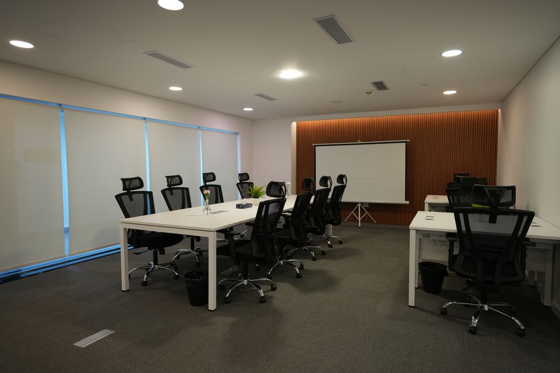 An empty meeting room with a long white table, black mesh chairs, a wood-panelled wall and a blank pull-down projector screen.