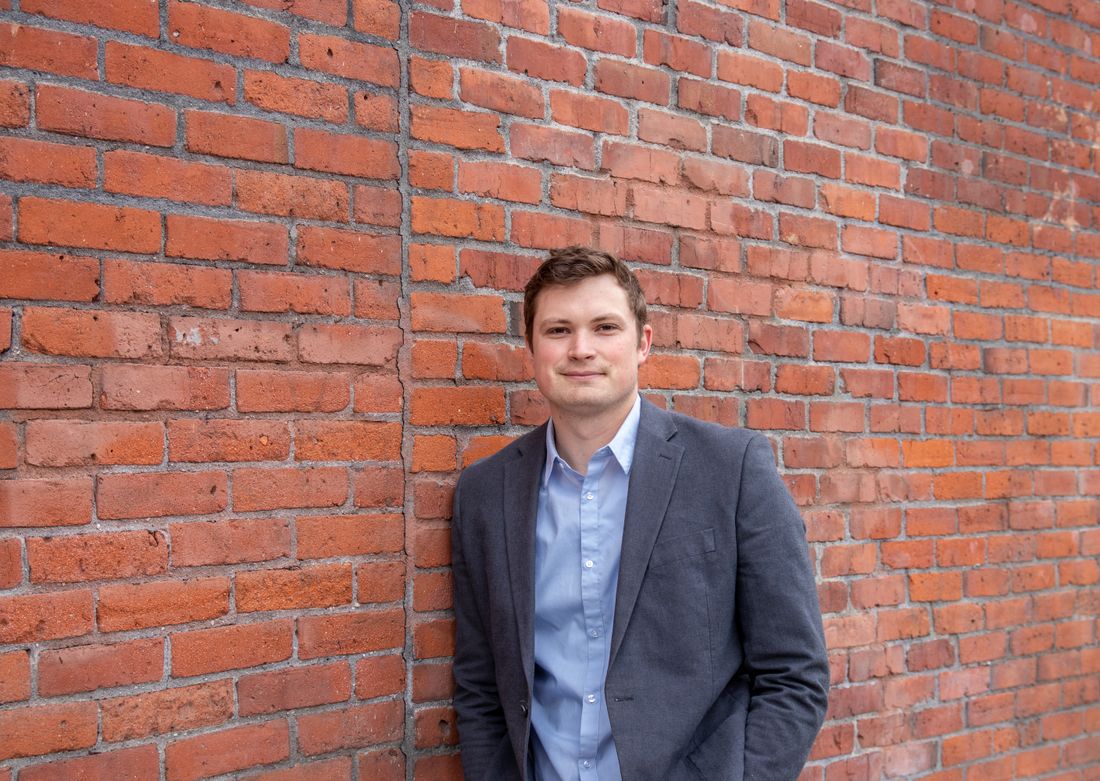 A man in a grey blazer and an open-collar blue shirt leaning against a red brick wall, smiling slightly.