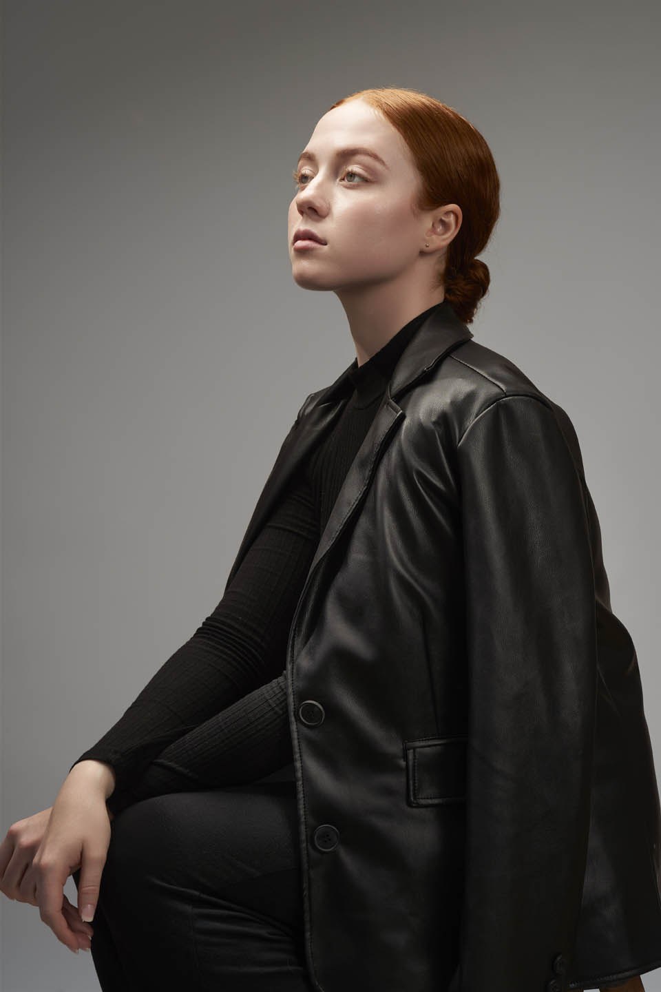 Woman in black leather blazer seated against grey background.