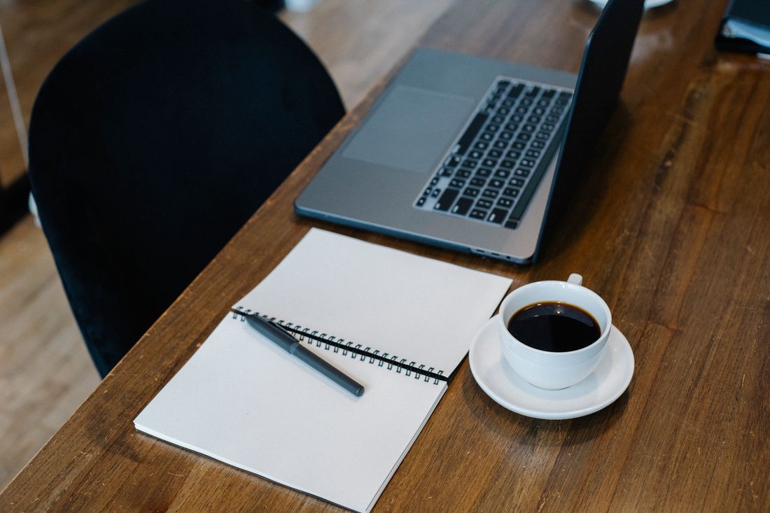 An open laptop, an open spiral notebook with blank pages, and a cup of coffee sit on a wooden table beside an empty chair.