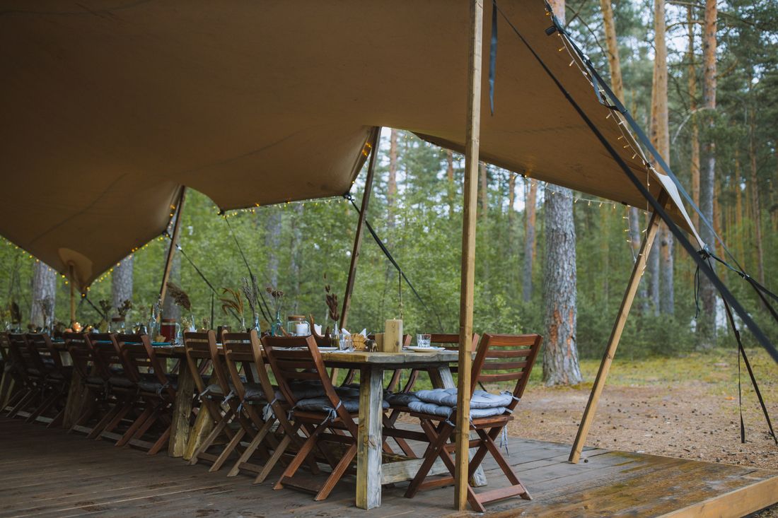 A view from beneath a canvas event tent's peaked roof and support poles, looking down a long dining table set with chairs in a wooded outdoor setting, no people present.