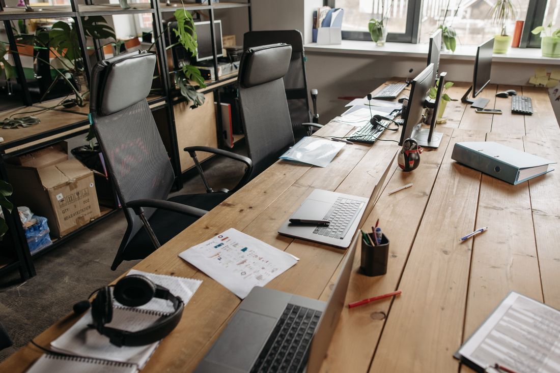 An empty modern co-working desk lined with laptops, monitors, and notebooks in a bright office space.