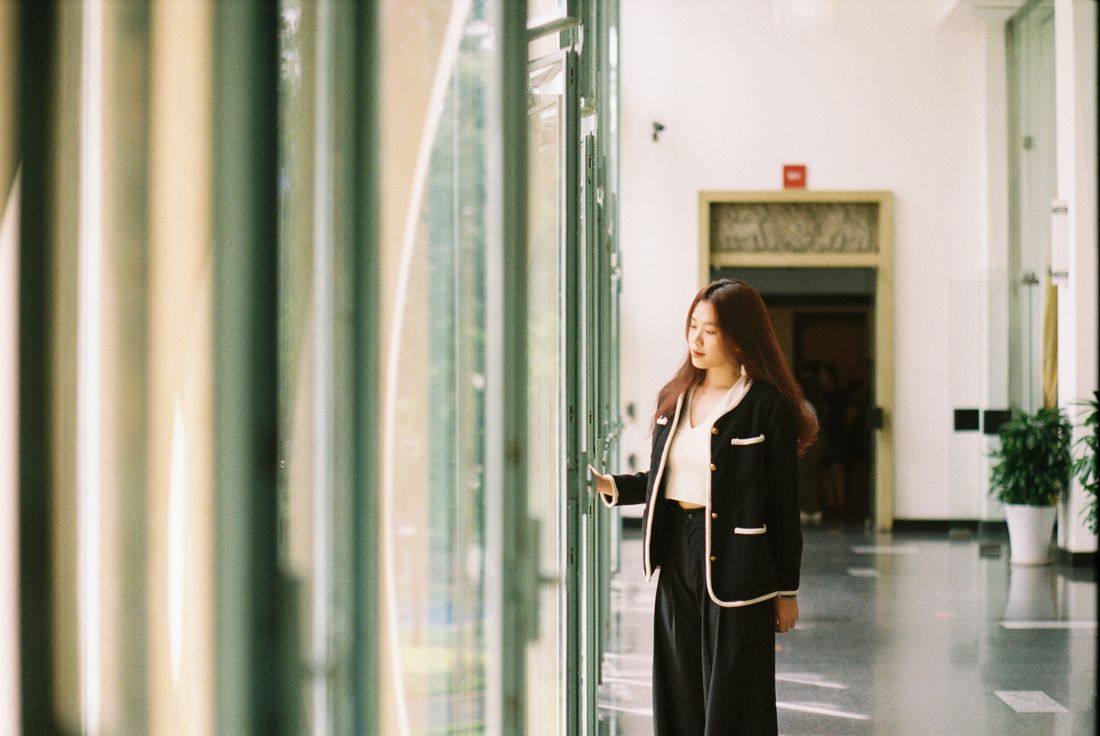 A woman in a tailored jacket stands near tall glass doors in a bright modern building, looking off to the side in natural daylight.