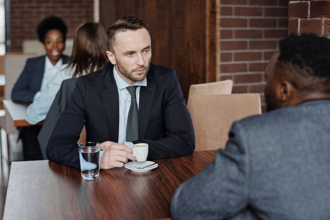 Two people in suits sit across a small wooden table in a cafe, leaning in over coffee and water in an unhurried conversation.