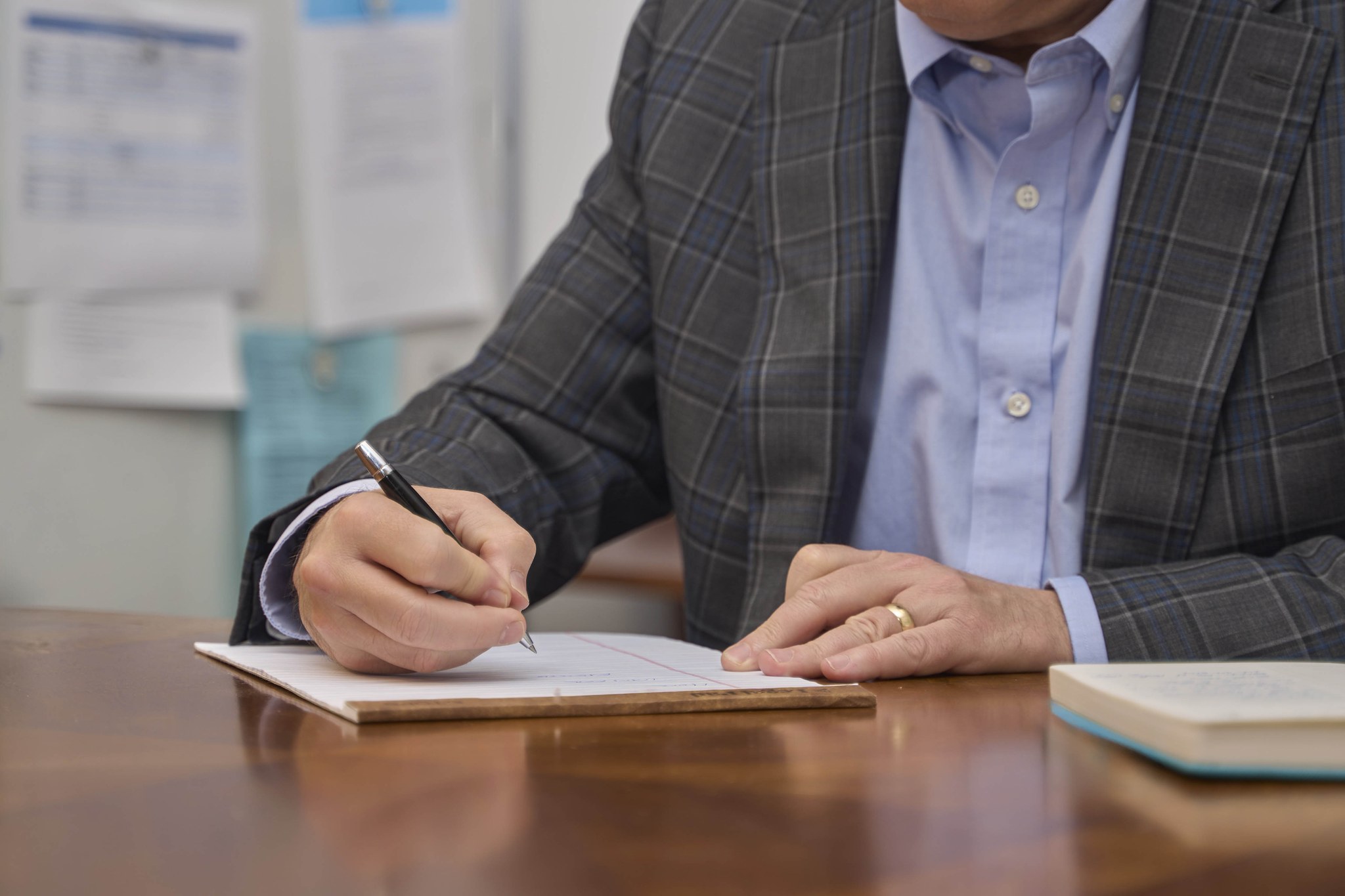A man's hand writes on a paper notepad with a pen at an office desk, his other hand resting on the page.